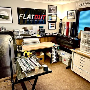 K&ouml;nnte beinhalten: Ein Arbeitsbereich mit einem gro&szlig;en schwarzen Banner mit dem Text FLATOUT GRAPHICS, einem Schreibtisch mit Glasplatte und kreisf&ouml;rmigen Mustern, einem wei&szlig;en Schubladenschrank, Vinylrollen, einem Gro&szlig;formatdrucker und Wandfelgen.