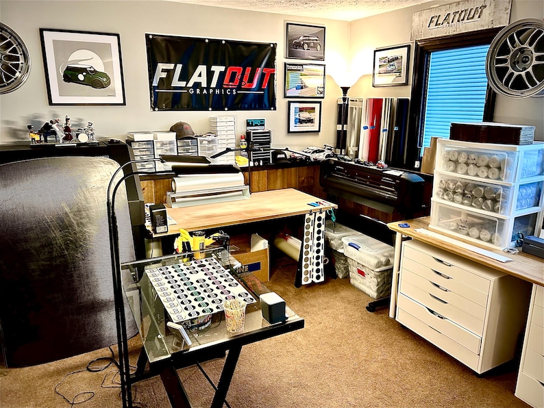 K&ouml;nnte beinhalten: Ein Arbeitsbereich mit einem gro&szlig;en schwarzen Banner mit dem Text FLATOUT GRAPHICS, einem Schreibtisch mit Glasplatte und kreisf&ouml;rmigen Mustern, einem wei&szlig;en Schubladenschrank, Vinylrollen, einem Gro&szlig;formatdrucker und Wandfelgen.
