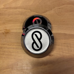 K&ouml;nnte beinhalten: Zwei kreisf&ouml;rmige Embleme mit metallisch silbernem Finish und erhabenem Rand. Das obere Emblem zeigt ein schwarzes Unendlichkeitssymbol auf silbernem Hintergrund. Das untere Emblem ist teilweise verdeckt und zeigt ein schwarzes Zentrum mit einem reflektierenden roten Unendlichkeitslogo.