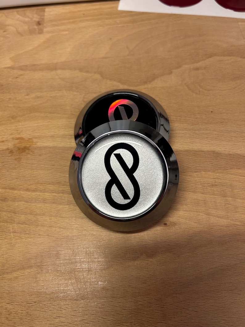 K&ouml;nnte beinhalten: Zwei kreisf&ouml;rmige Embleme mit metallisch silbernem Finish und erhabenem Rand. Das obere Emblem zeigt ein schwarzes Unendlichkeitssymbol auf silbernem Hintergrund. Das untere Emblem ist teilweise verdeckt und zeigt ein schwarzes Zentrum mit einem reflektierenden roten Unendlichkeitslogo.