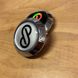 K&ouml;nnte beinhalten: Zwei kreisf&ouml;rmige Radnabendeckel mit Chrom-Finish. Der vordere Deckel hat einen silbernen metallischen Hintergrund mit einem schwarzen Unendlichkeitssymbol-Logo. Der teilweise sichtbare hintere Deckel hat einen schwarzen Hintergrund mit einem bunten, irisierenden Logo.