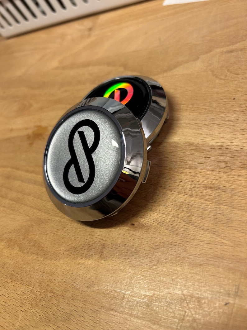 K&ouml;nnte beinhalten: Zwei kreisf&ouml;rmige Radnabendeckel mit Chrom-Finish. Der vordere Deckel hat einen silbernen metallischen Hintergrund mit einem schwarzen Unendlichkeitssymbol-Logo. Der teilweise sichtbare hintere Deckel hat einen schwarzen Hintergrund mit einem bunten, irisierenden Logo.