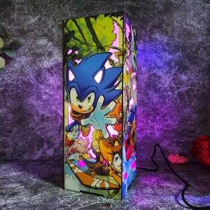 Könnte beinhalten: Eine rechteckige Holzlampe mit einem lebendigen Grafikdruck von Sonic the Hedgehog und anderen Charakteren aus dem Franchise. Die Lampe strahlt ein helles violettes und rosa Licht aus, das die detaillierten Cartoon-Grafiken auf der Oberfläche beleuchtet.