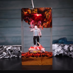 Könnte beinhalten: Eine rechteckige Kunstharzlampe mit einer Figur mit Hockeymaske, die eine Axt hält und auf einem Sockel mit rot-orangen Lavatexturen steht. Der obere Teil enthält ein rotes Licht und auf dem Sockel ist der Schriftzug Friday 13th zu sehen.