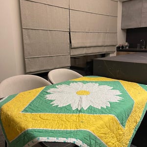 May include: A green and yellow quilted table runner with a white daisy in the center. The runner has a scalloped edge and is made of cotton fabric.