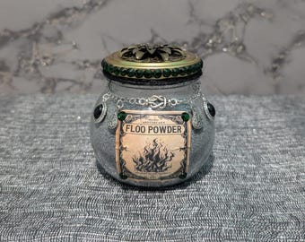 Op de afbeelding: Een decoratieve apothekerspot met een zilvergrijze buitenkant, versierd met een bronzen deksel en groene accenten. De pot heeft een label met de tekst "FLOO POWDER" met een vlammenafbeelding. Een zilveren ketting met zwarte en groene versieringen versiert de pot.