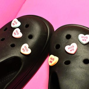 May include: Black clogs with heart-shaped charms that read "HUG ME" in white, pink, and yellow. The charms are placed on the top of the clogs. The background is a solid pink color.