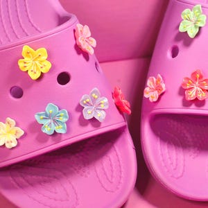 May include: A pair of bright pink slide sandals adorned with colorful flower charms. The sandals have multiple holes and a textured footbed. The flower charms are in shades of yellow, pink, blue, and green.