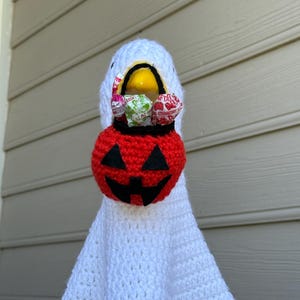 Pode incluir: Uma fantasia de fantasma de croch&ecirc; branco feita &agrave; m&atilde;o com uma cesta de doces de ab&oacute;bora vermelha. A cabe&ccedil;a do fantasma &eacute; branca com um bico amarelo e segura uma cesta de ab&oacute;bora vermelha e preta cheia de doces embalados, perfeita para o Halloween.