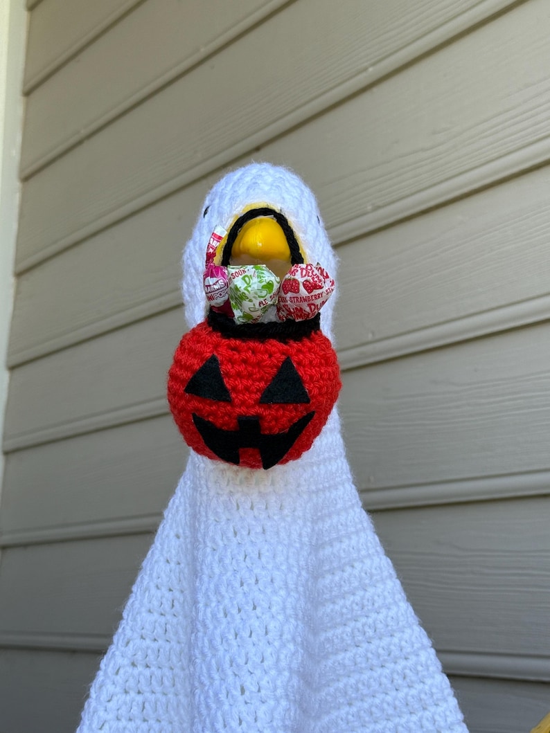 Pode incluir: Uma fantasia de fantasma de croch&ecirc; branco feita &agrave; m&atilde;o com uma cesta de doces de ab&oacute;bora vermelha. A cabe&ccedil;a do fantasma &eacute; branca com um bico amarelo e segura uma cesta de ab&oacute;bora vermelha e preta cheia de doces embalados, perfeita para o Halloween.