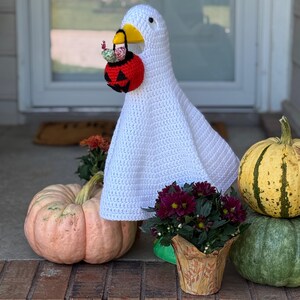 Pode incluir: Um ganso branco de croch&ecirc; usando uma fantasia de Halloween, segurando um balde de doces de ab&oacute;bora vermelha. O ganso est&aacute; rodeado por ab&oacute;boras e flores em vasos, criando uma exibi&ccedil;&atilde;o de outono.