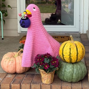 Pode incluir: Uma decora&ccedil;&atilde;o de ganso rosa de croch&ecirc; com um bico amarelo e uma bolsa roxa no bico, cheia de flores. Duas ab&oacute;boras, uma laranja e outra verde, e uma ab&oacute;bora listrada amarela e verde est&atilde;o por perto. Um vaso de flores roxas tamb&eacute;m est&aacute; presente.