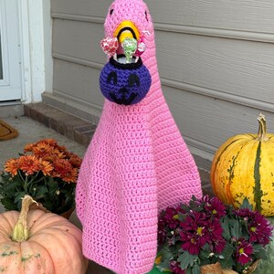 Pode incluir: Um ganso rosa de croch&ecirc; usando um porta-doces de ab&oacute;bora roxa. O ganso est&aacute; rodeado por ab&oacute;boras e cris&acirc;ntemos em vasos, criando uma exibi&ccedil;&atilde;o festiva de Halloween. O porta-doces est&aacute; cheio de pirulitos coloridos.