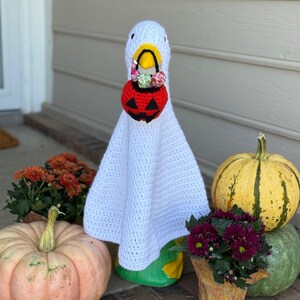 Pode incluir: Um ganso branco de croch&ecirc; usando uma fantasia de fantasma branca e segurando uma cesta de doces em forma de ab&oacute;bora vermelha. A cena inclui ab&oacute;boras e flores em vasos, criando uma decora&ccedil;&atilde;o de Halloween.