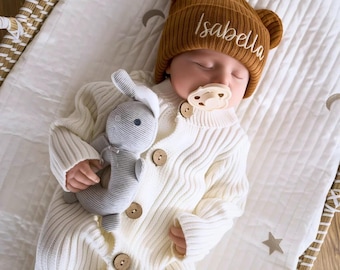 Pode incluir: Um bebê vestindo um cardigã de tricô branco e um gorro de tricô marrom com o nome "Isabella" bordado. O bebê está segurando um brinquedo de coelho de pelúcia cinza e está usando uma chupeta.