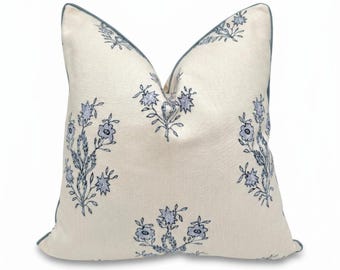 May include: A square throw pillow in an off-white fabric featuring a repeating blue floral block print pattern. The pillow is finished with a solid blue piped edge trim that outlines the square shape, providing a clean and defined border.