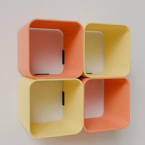 May include: Four square, open-faced shelves in a stacked arrangement. Two are peach-colored, and two are yellow. Each shelf has a white interior and a small black accent. The shelves are mounted on a white wall.