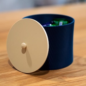 May include: A navy blue cylindrical container with a cream-colored lid. The lid is partially open, revealing the contents inside. The container has a ribbed texture and a small knob on the lid. The container is sitting on a wooden surface.