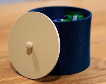 以下が含まれることがあります： クリーム色の蓋が付いた、ネイビーブルーの円筒形の容器。 蓋は部分的に開いており、中身が見えます。 容器はリブ加工が施されており、蓋には小さなノブが付いています。 容器は木製の表面に置かれています。