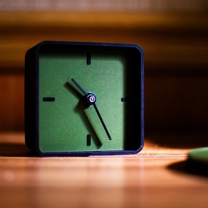 May include: A square, black-framed clock with a green face. The clock hands point to approximately 5:25. The clock sits on a wooden surface, with a blurred green object in the foreground. The background is a wooden wall.