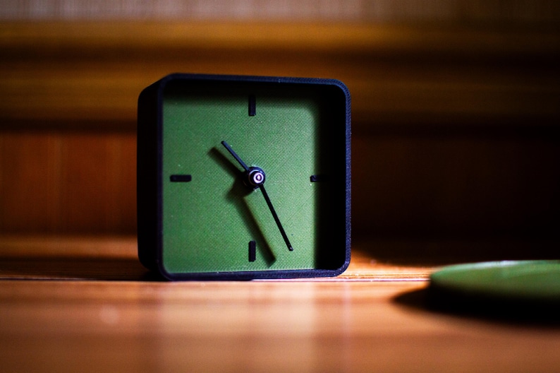 May include: A square, black-framed clock with a green face. The clock hands point to approximately 5:25. The clock sits on a wooden surface, with a blurred green object in the foreground. The background is a wooden wall.