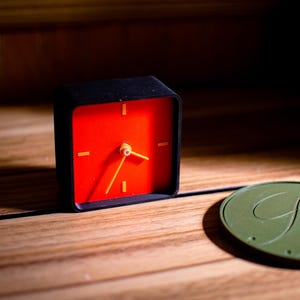 May include: A small, square black alarm clock with a bright red face and yellow hands and markers. The clock sits on a wooden surface next to a green, circular object with the letters "GM" in a cursive font.