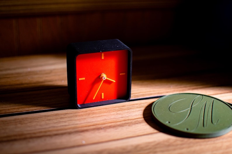 May include: A small, square black alarm clock with a bright red face and yellow hands and markers. The clock sits on a wooden surface next to a green, circular object with the letters "GM" in a cursive font.