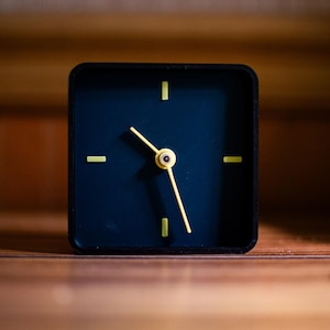 Puede incluir: Un reloj analógico negro cuadrado con una esfera azul oscuro y manecillas y marcadores amarillos. El reloj está sobre una superficie de madera. El fondo es una pared de madera borrosa.