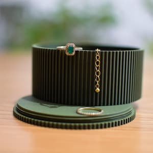 May include: A green ring holder with a ribbed texture. The holder has a ring with a green stone and a gold chain hanging from it. There is a second ring on the bottom of the holder.