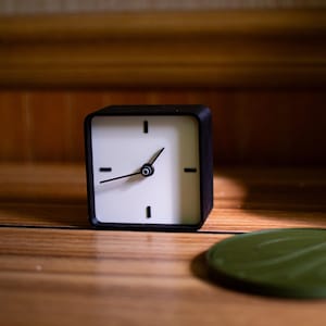May include: A square black and white analog clock with black hands and a green circular object on a wooden surface. The clock has a minimalist design with black markings for the hours.