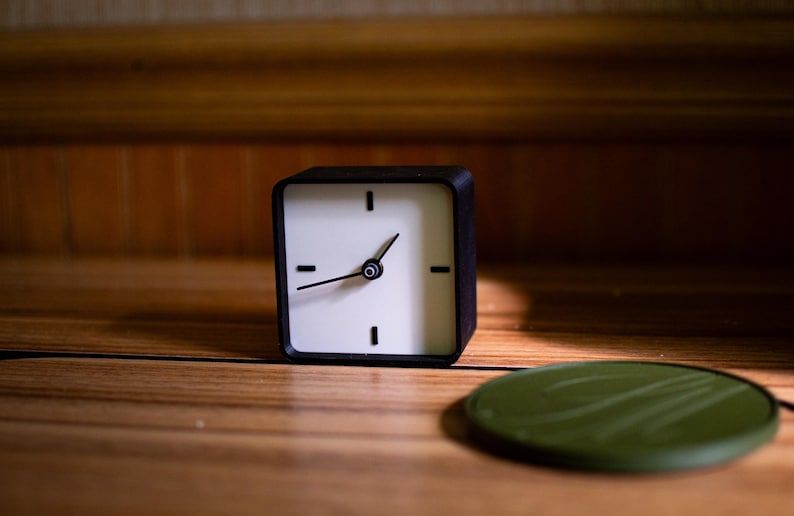 May include: A square black and white analog clock with black hands and a green circular object on a wooden surface. The clock has a minimalist design with black markings for the hours.
