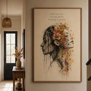 May include: A rectangular wall art print featuring a black ink sketch of a profile face with watercolor flowers in shades of orange, pink, and blue. The text "who are you losing while finding yourself" is printed in black at the top of the cream background.