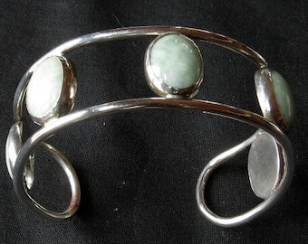 May include: Silver bangle bracelet with three oval-shaped green gemstones set in the metal. The bracelet has a hinged opening.