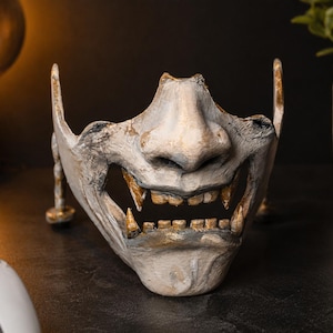 Könnte beinhalten: Eine weiß-graue Halbmaske mit einer modellierten Nase, markanten Fangzähnen und sichtbaren Zähnen. Die Oberfläche weist ein verwittertes, strukturiertes Finish mit dezenten goldenen Akzenten an den Kanten und Gesichtszügen auf.