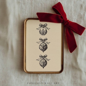 May include: A framed print featuring three detailed black and white Christmas ornament illustrations with bows. The frame has a gold finish, and a red velvet bow is tied on the top right. The text "FOXGLOVE WALL ART" is at the bottom.