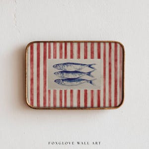 May include: Rectangular wall art featuring three blue fish illustrations on a cream background, set against a red and white striped pattern. The artwork is framed with a gold-colored metal edge. The text "FOXGLOVE WALL ART" is at the bottom.