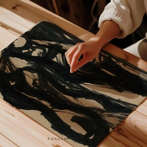 May include: A close-up of a piece of art with bold black brushstrokes on a beige paper. The artwork is on a wooden surface. The artist's hand is visible, adding a human element to the abstract composition. The word "FOXGLOVE" is printed at the bottom left.