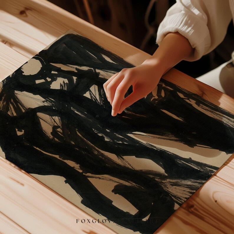 May include: A close-up of a piece of art with bold black brushstrokes on a beige paper. The artwork is on a wooden surface. The artist's hand is visible, adding a human element to the abstract composition. The word "FOXGLOVE" is printed at the bottom left.