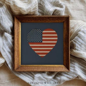 May include: Framed wall art featuring a heart design with the American flag. The heart is divided into the stars and stripes of the flag, set against a navy blue background. The frame is a warm, dark brown color.