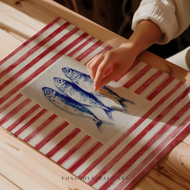 May include: A watercolor painting of four blue fish on a textured white paper, set against a red and white striped background. The artwork is on a wooden surface. The artist's hand is touching the artwork. The text "FOXGLOVE WALL ART" is at the bottom.