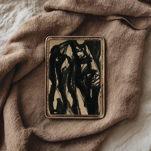 May include: A rectangular framed artwork with an abstract black ink design on a beige background. The frame is gold-colored, and the artwork is displayed on a brown fabric surface. The artwork is a modern piece.