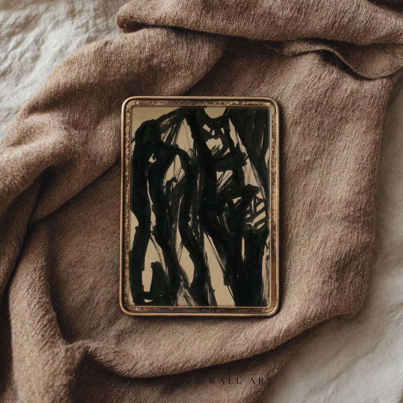 May include: A rectangular framed artwork with an abstract black ink design on a beige background. The frame is gold-colored, and the artwork is displayed on a brown fabric surface. The artwork is a modern piece.