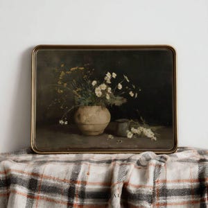 May include: A framed still life painting featuring a beige vase filled with white and yellow flowers. A small, dark gray container and a small bouquet of white flowers are also visible. The painting has a dark background and a gold frame.