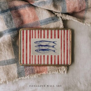 May include: Rectangular wall art with a gold frame. The artwork features a red and white striped background with three blue fish illustrations. The text "FOXGLOVE WALL ART" is at the bottom. The art is set on a textured fabric.
