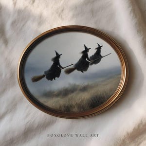 May include: Oval framed wall art featuring three witches flying on brooms against a muted landscape. The witches wear black dresses and pointed hats. The frame is gold-colored, and the artwork is set against a neutral background. The text "FOXGLOVE WALL ART" is at the bottom.