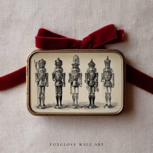 May include: A rectangular, gold-framed print featuring five detailed illustrations of nutcrackers. The print is tied with a deep red velvet ribbon. The text "FOXGLOVE WALL ART" is printed at the bottom of the frame.