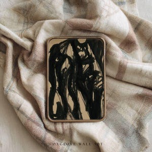 May include: A rectangular framed artwork with a gold-colored border. The artwork features a black abstract design on a beige background. The artwork is placed on a beige, brown, and blue plaid blanket. The text "FOXGLOVE WALL ART" is visible at the bottom.