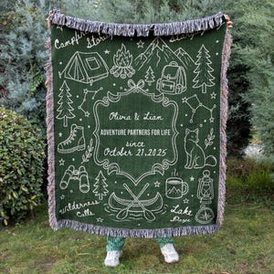 May include: A dark green woven throw blanket with fringed edges featuring white line art of camping gear, mountains, trees, a fox, and a canoe. The center text reads: Olivia & Liam, ADVENTURE PARTNERS FOR LIFE, since October 21, 2025.
