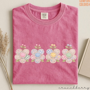 May include: A pink Comfort Colors t-shirt with a floral design. The design features four appliqued flowers with gingham and floral patterns in the center. The shirt has a crew neck and the text "crunchberry" is at the bottom.