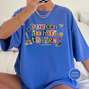 May include: A bright blue t-shirt featuring the text "you can do hard things" in a colorful, multi-font collage style. The design includes small graphic illustrations of a pencil, a rainbow, a paper airplane, a smiley face, a heart, and a sneaker.